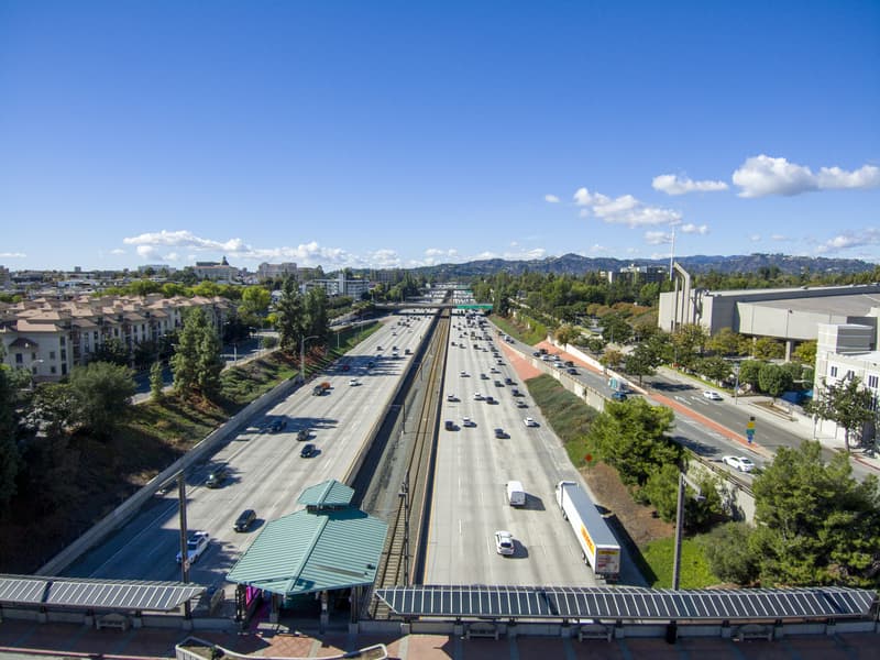Aerial shot of cars and trucks driving along the 210 Freeway surrounded by lush green trees and plants, apartments